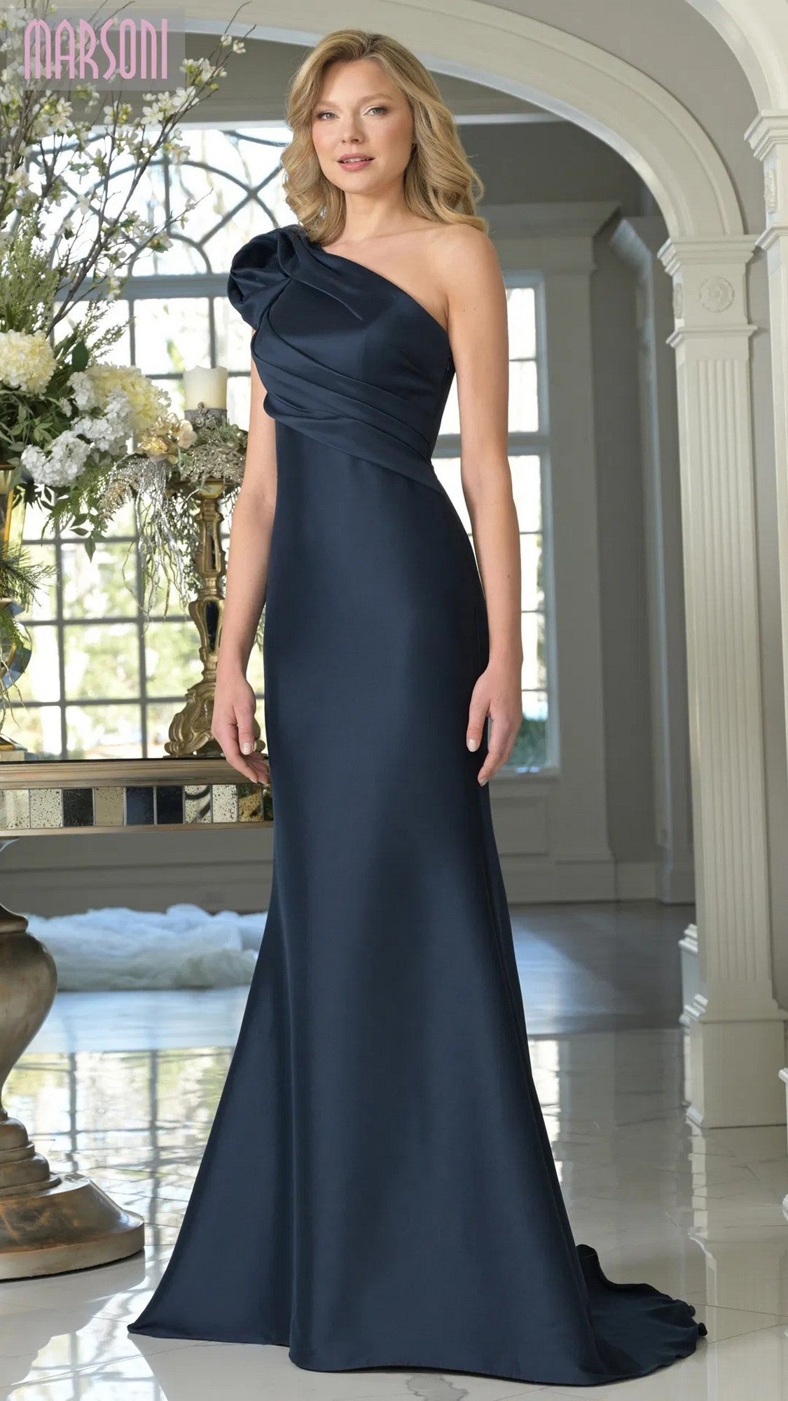 A woman wearing the Marsoni MV1358 long formal dress in navy stands indoors on a marble floor near floral arrangements and arched windows.